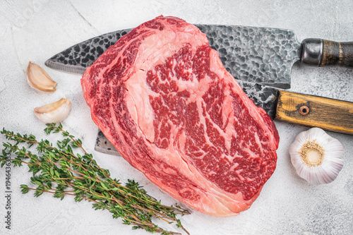 Premium raw ribeye beef steak with intense marbling and thick fat cap. Rustic butcher style on light stone background.