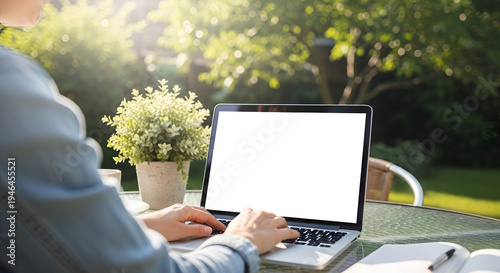 Person working outdoors on laptop with white screen surrounded by lush green garden using for remote work and online communication