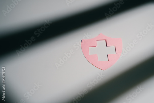 Healthcare concept. Pink shield with cross cutout on white surface with shadow lines, symbolizing protection and healthcare in calm setting