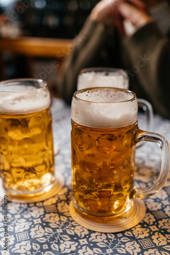 Close-up of beer mugs with thick foam, as a woman holds one glass and brings it to her lips. The image conveys relaxation, social drinking, and a cozy bar atmosphere.