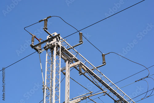 A complex steel lattice structure supports high voltage overhead power lines for a railway system against a clear blue sky