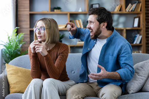 Man angrily pointing and yelling at a frightened woman on a couch, tense domestic dispute showing verbal abuse, fear, and relationship conflict in a home setting