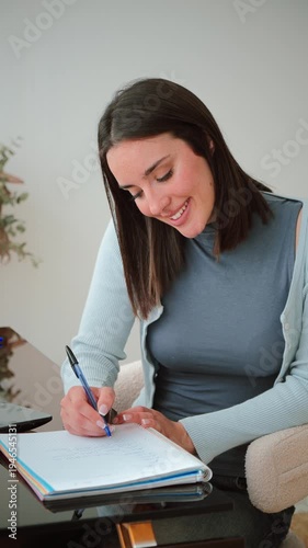 Engaged woman thoughtfully writing notes in her notebook, showcasing concentration and creativity during her work session at a stylish desk with a pen in hand and a smile on her face.