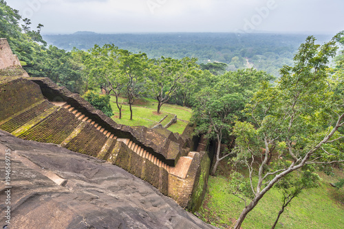 Sigiriya ancient rock fortress in Sri Lanka. Ancient Rock Fortress Staircase Descend to Lush Green Gardens and Expansive Forest Canopy Under Overcast Sky