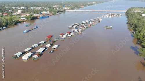 Aerial drone view of Chau Doc floating village along Hau River showing rows of houses boats fish farms across muddy Mekong water near An Giang with bridge and riverside settlement