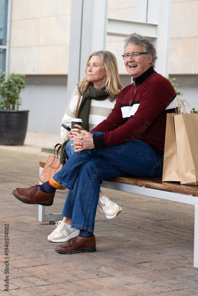 Fototapeta premium Senior couple wearing sweaters and jeans sitting on wooden bench in plaza with coffee, paper bags