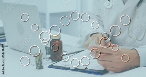 Doctor wearing white lab coat, stethoscope holding pill bottle, checking labels at desk with laptop
