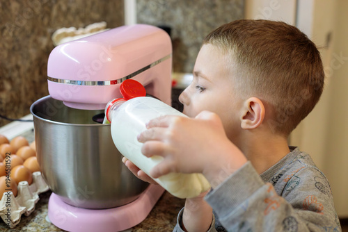 little boy pouring milk into stand mixer bowl while cooking pancake batter in kitchen, child helping prepare homemade breakfast dough with eggs and ingredients