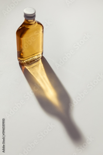 Small glass bottle with amber liquid casting a long, glowing golden shadow on a white surface. Elegant product photography with bright sunlight, caustic light effects, and minimalist aesthetic.