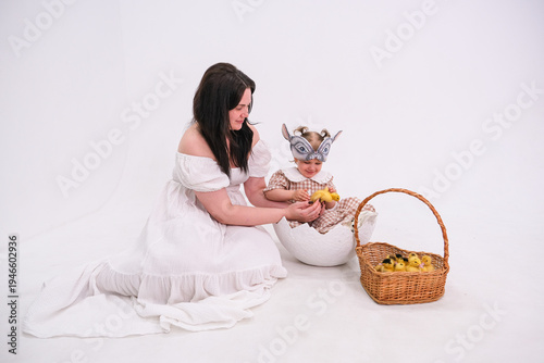 Moscow, Russia, 12.04.2025 Woman in white dress and little child in bunny mask play with small yellow ducklings. Easter photoshoot celebration with basket and decorative broken eggshell. Spring family