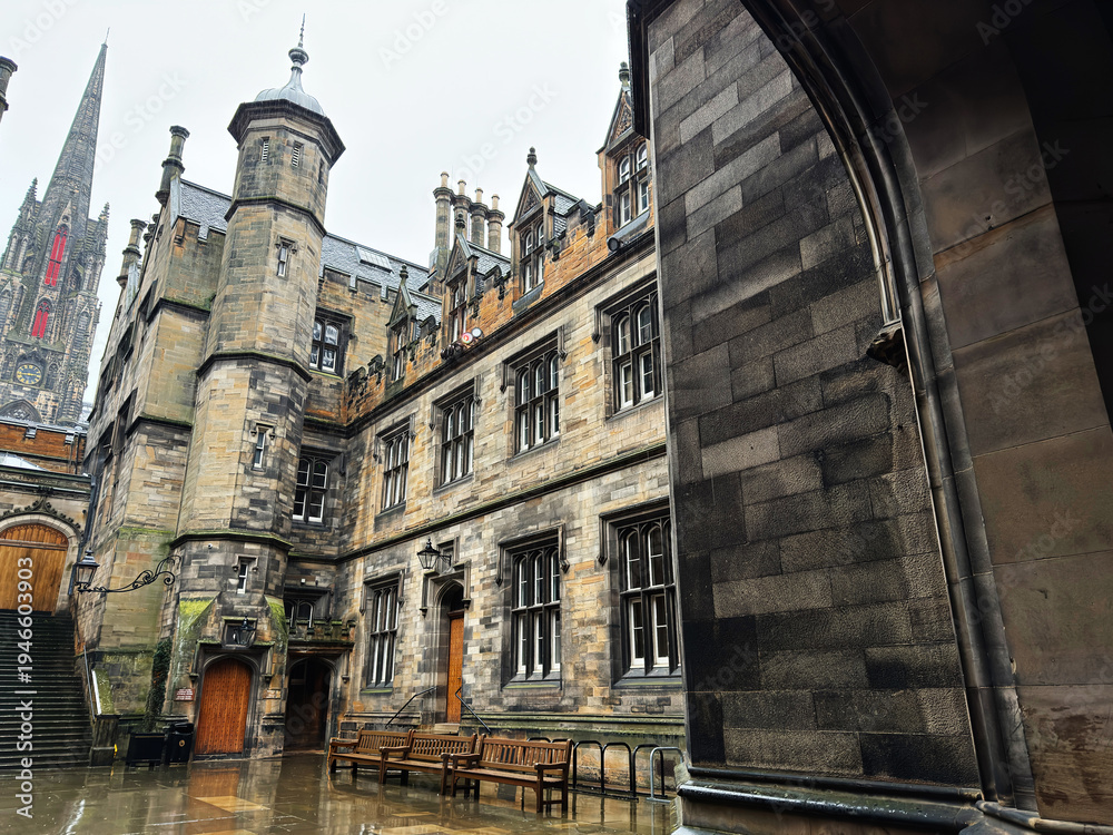 Fototapeta premium Historic stone New College Quad courtyard and gothic spire in Edinburgh, Scotland, UK with wet pavements, benches and ornate university style architecture