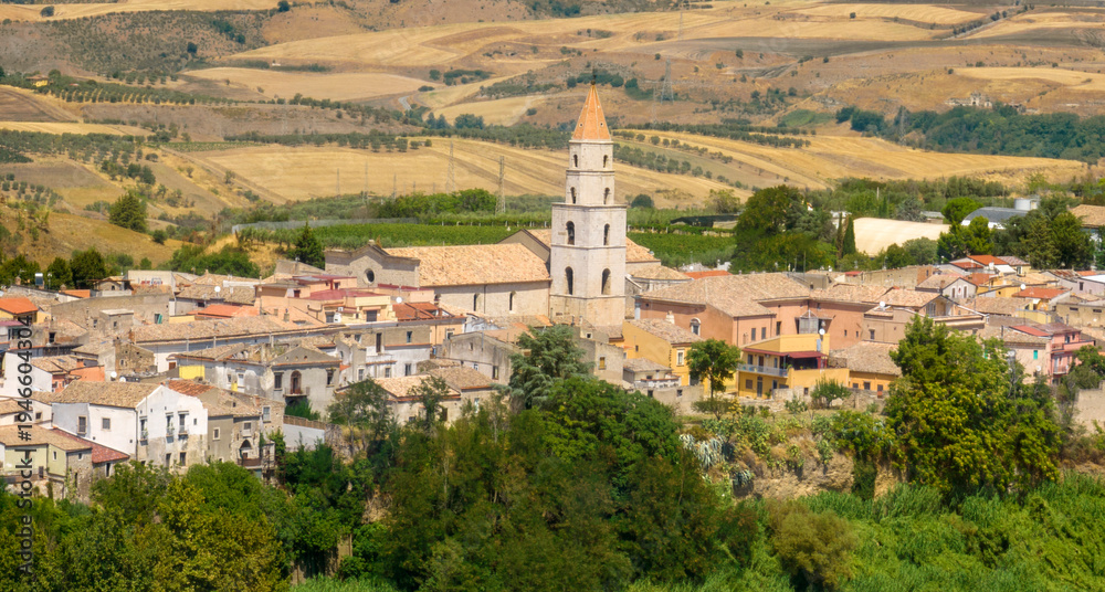 Fototapeta premium Aerial view of a traditional Italian village nestled in rolling hills. A prominent white bell tower with a pointed orange roof rises above the town's historic buildings. It is Venosa, in Basilicata.