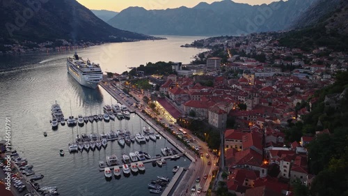 Aerial establishing view of Kotor Old town lights in blue hour, mountains range of Kotor Bay fjord in a background, large cruise ship in the port. 