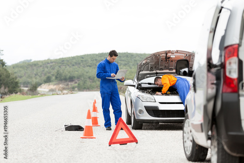 Obraz na płótnie Two professional mechanics in colorful uniforms repairing a broken car on the roadside with orange traffic cones for safety