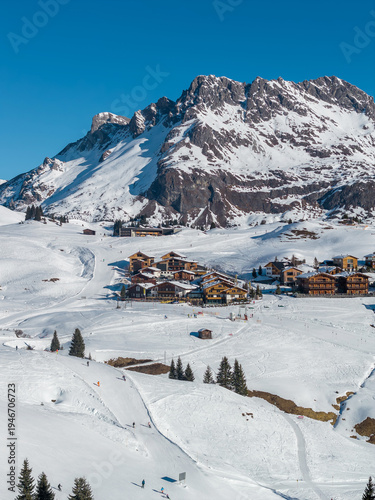 Aerial winter scene of Lech and Oberlech in the Austrian Alps shows chalets, slope side hotels, groomed pistes, ski lifts, resting skiers, evergreens, and a jagged peak.