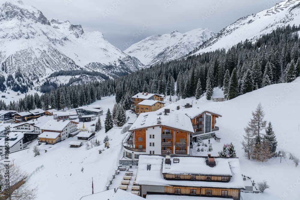 Fototapeta premium Aerial winter view of Lech and Oberlech in the Austrian Alps shows chalets, luxury hotels, groomed pistes, winding roads, and spruce forests under soft overcast light.