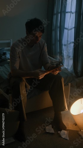 Vertical shot of depressed Caucasian man taking old photograph from notebook and tearing it apart while sitting on bed by flickering lamplight at nighttime