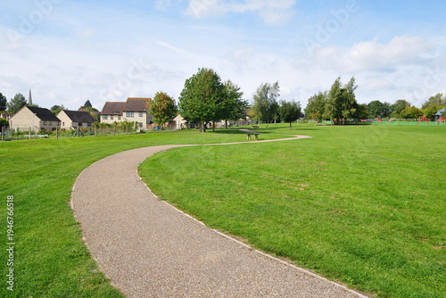 A winding gravel footpath meanders through a vibrant green park with lush grass recreation field, leafy trees and blue sky above