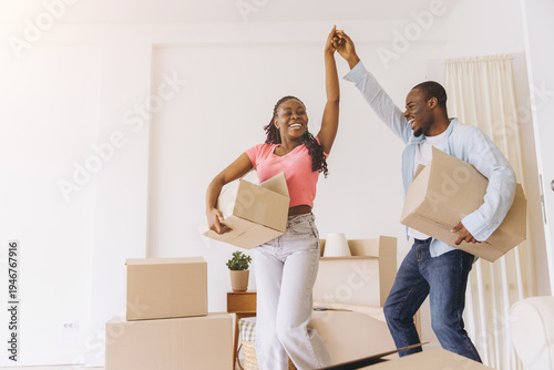 Happy young black couple celebrating moving day and new home, dancing and enjoying relocation with cardboard boxes