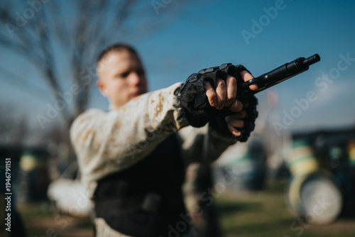 Tableau sur toile Man wearing gloves and a vest points a paintball gun forward in an outdoor field