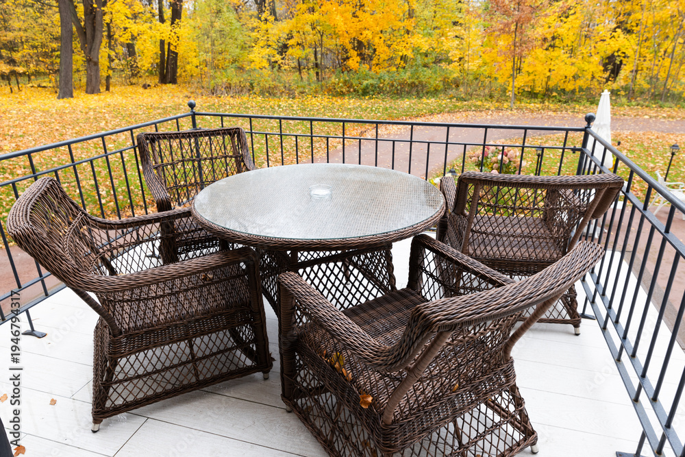 Fototapeta premium Wicker patio table and chairs arranged on a balcony with vibrant yellow autumn trees beyond, suggesting cozy outdoor dining, relaxation and a peaceful seasonal atmosphere