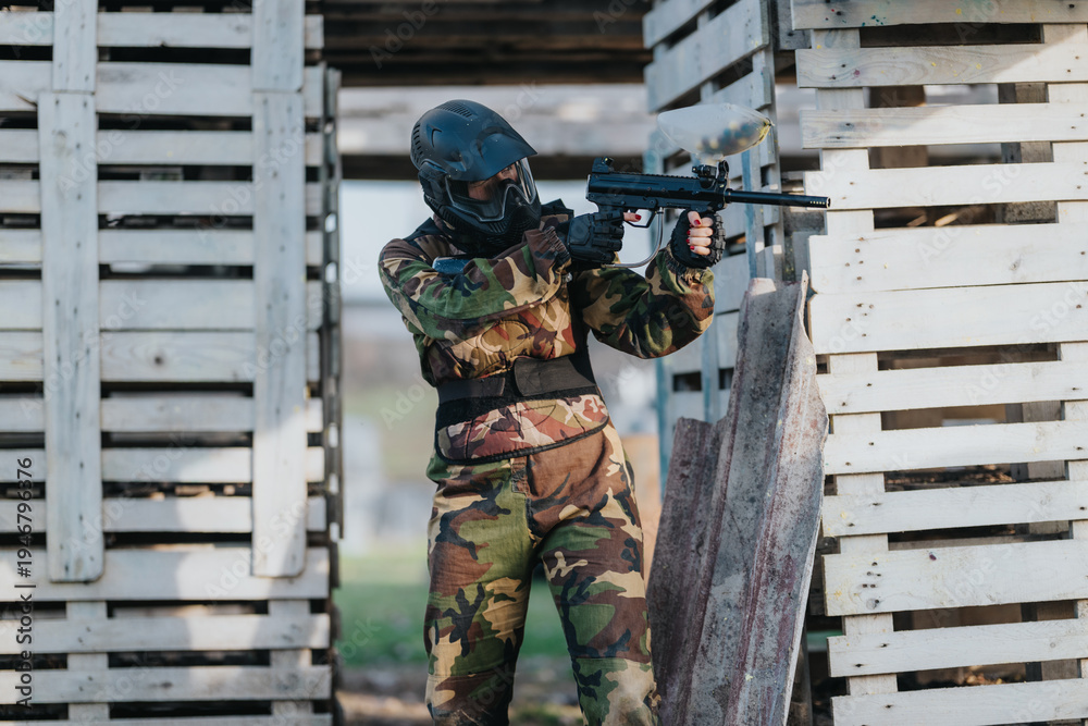Fototapeta premium A masked airsoft player in camouflage holds a replica rifle behind a wooden bunker. The scene captures a tense moment in an outdoor airsoft game.