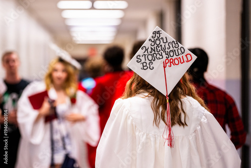 Photography An unrecognizable teenager wearing a white graduation gown walks down a school hallway, their decorated mortarboard bearing the powerful message If you knew my story, you would be proud