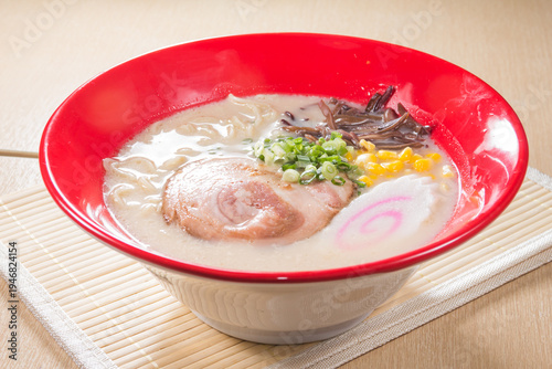 A red rim bowl holds creamy tonkotsu ramen with chashu, corn, scallion, wood ear, and a pink swirl fish cake. The rich broth and steam suggest classic Japanese comfort food.