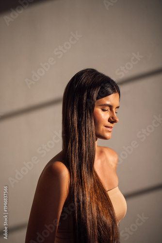 Young woman with long dark hair stands in soft light, eyes closed, showcasing serene expression and natural beauty, with shadows creating a calming atmosphere