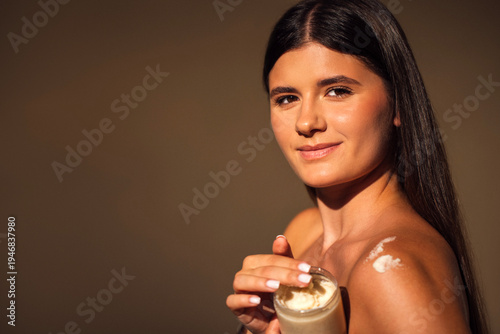 Young woman with long dark hair holds a jar of cream, applying skincare product to her shoulder, showcasing beauty routine and self-care in a warm, inviting atmosphere