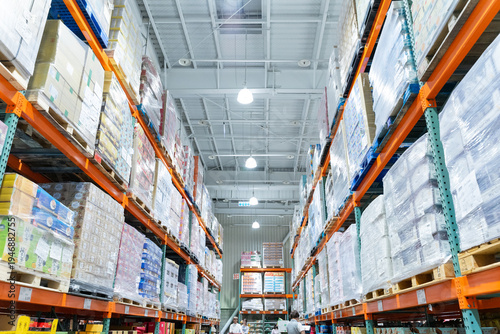 A wide interior photograph of a large warehouse-style wholesale shopping center featuring long aisles lined with tall pallet racks filled with bulk merchandise. The image captures the spacious retail 