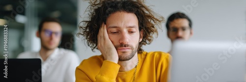 Male caucasian young adult in yellow sweater leaning on hand looking bored at laptop in classroom or office with colleagues blurred behind him conveying distraction and disengagement