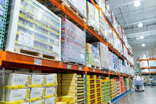 A wide interior photograph of a large warehouse-style wholesale shopping center featuring long aisles lined with tall pallet racks filled with bulk merchandise. The image captures the spacious retail 