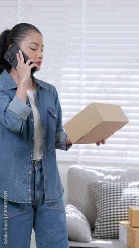 Asian woman small business owner standing still holding parcel box while talking slowly on smartphone and reviewing delivery information online shopping customer service routine inside home workspace