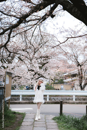 Young woman in white coat and boots standing on stone path under blooming cherry blossom tree in urban park during spring