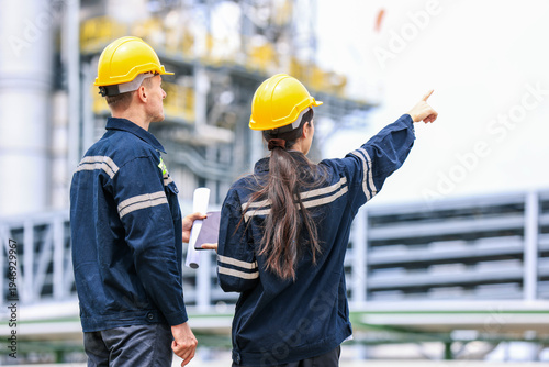 group of industrial engineers workers in a refinery - oil and gas processing equipment and machinery, engineers collaborate with a laptop, blueprint, and digital tablet at the oil storage tanks site.