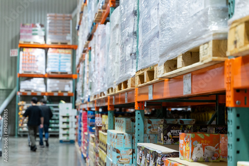  A wide interior photograph of a large warehouse-style wholesale shopping center featuring long aisles lined with tall pallet racks filled with bulk merchandise. The image captures the spacious retail