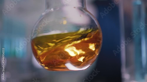 Female researcher preparing a new type of enriched bio fuel. Holding a glass flask with golden liquid close to the camera and slowly swirling the liquid. Selective focus