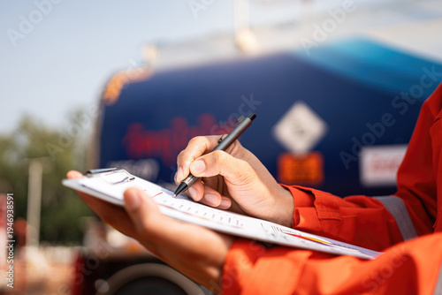 An engineer is checking on chemical hazardous material checklist to verify the safety condition with chemical road tanker as background. Industrial waorking scene, close-up and selective focus.