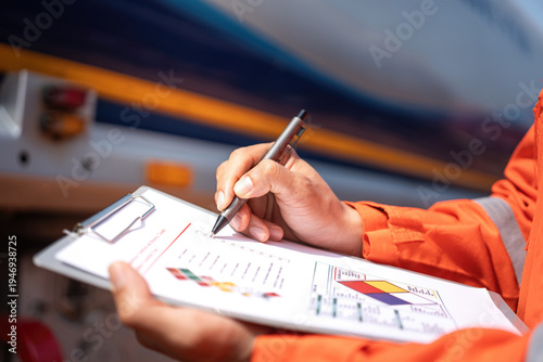 An engineer is checking on chemical hazardous material checklist to verify the safety condition with chemical road tanker as background. Industrial waorking scene, close-up and selective focus.