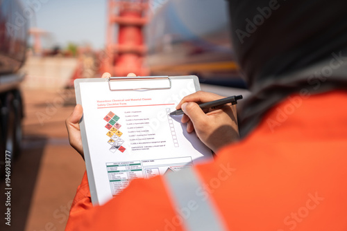 An engineer is checking on chemical hazardous material checklist to verify the safety condition with chemical road tanker as background. Industrial waorking scene, close-up and selective focus.