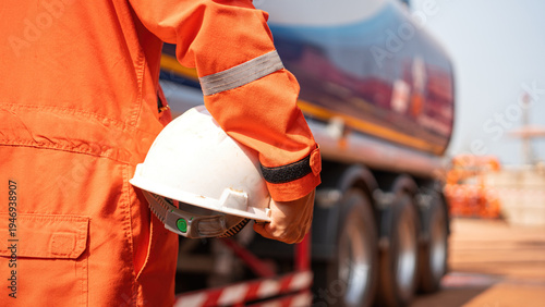 A worker in orange coverall uniform is holding a white safety helmet with background from road tanker truck. Ready to working in risk workplace, close-up and selective focus.