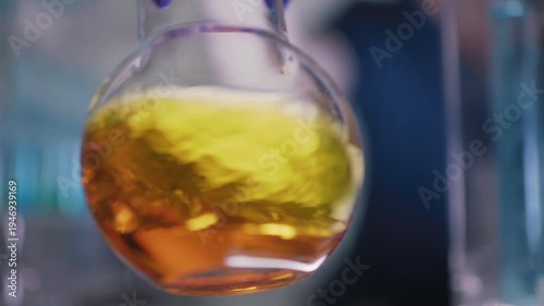 Female researcher preparing a new type of enriched fuel. Holding a glass flask with golden liquid close to the camera and swirling the liquid. Selective focus