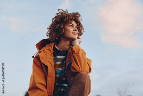Konstfotografi A smiling woman with curly hair wearing a rainbow sweater and orange jacket looks thoughtfully into the distance outdoors
