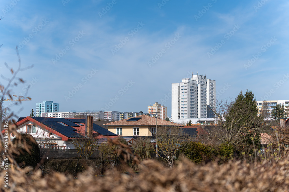 Fototapeta premium Clear blue sky above city skyline. Modern buildings stand behind residential rooftops. Trees and dry grass frame the foreground. Urban landscape viewed from a distance
