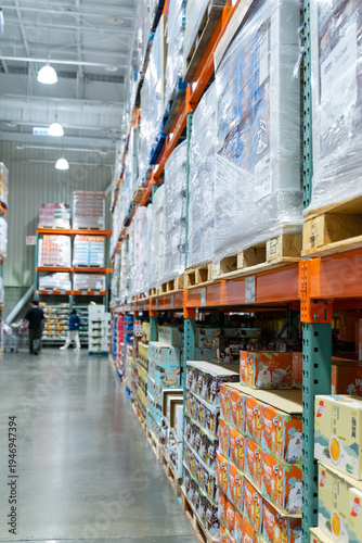 Photography A wide interior photograph of a large warehouse-style wholesale shopping center featuring long aisles lined with tall pallet racks filled with bulk merchandise