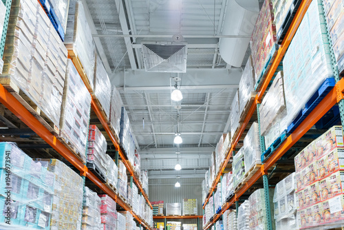  A wide interior photograph of a large warehouse-style wholesale shopping center featuring long aisles lined with tall pallet racks filled with bulk merchandise. The image captures the spacious retail