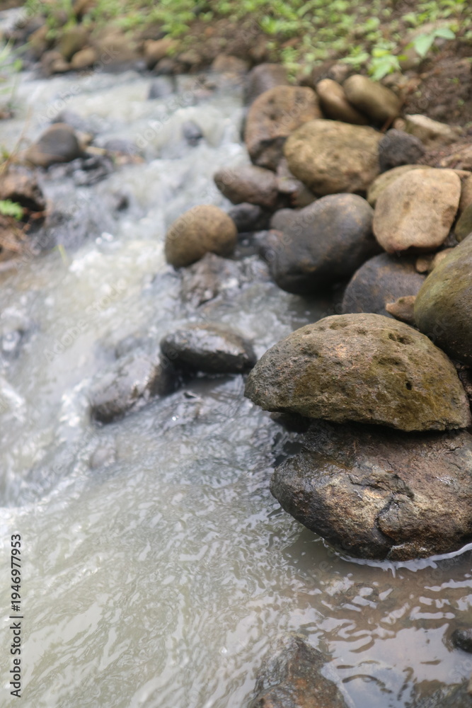 Obraz premium Turbid water flowing rapidly through a rocky riverbed surrounded by natural vegetation.
