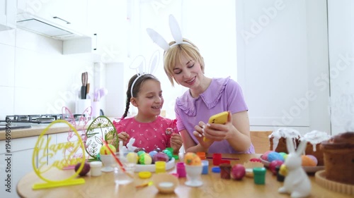 A mother and her daughter wearing bunny ears are decorating Easter eggs together in a bright kitchen while looking at a phone for inspiration