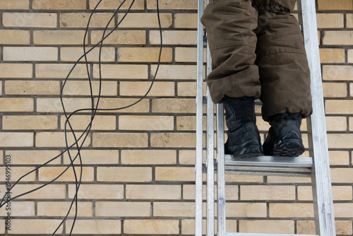 Worker's legs in brown pants and black boots on a metal ladder against a beige brick wall. Black cables hang nearby, suggesting construction or repair. Industrial feel with ample copy space
